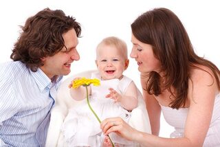 Das Bild zeit Eltern mit ihrem Kleinkind. Die Eltern halten dem Kind eine gelbe Blume hin und es freut sich.