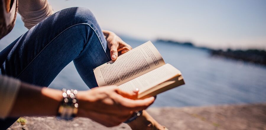 Auf dem Foto ist eine Frau zu erkennen, die ein Buch  liest. Sie sitzt auf einer Mauer, im Hintergrund erkennt man einen See oder auch das Meer.
