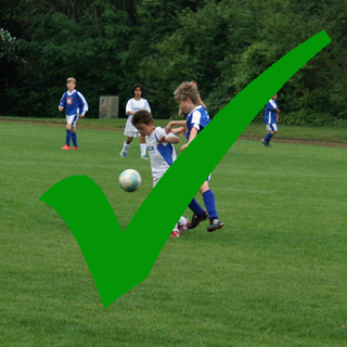 Das Symbolbild zeigt junge Fußballer*innen beim Fußballspiel auf einem Rasenplatz in Frechen - hier im Sportpark Herbertskaul. Im Bild ist ein grüner Haken, dieser symbolisiert, dass der Platz bespielbar ist.