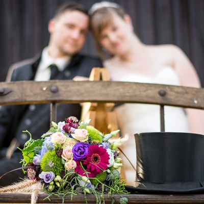 Das Bild zeigt einen Blumenstrauß und einen Hut-Zylinder. Im Hintergrund ist ein Brautpaar zu erkennen.