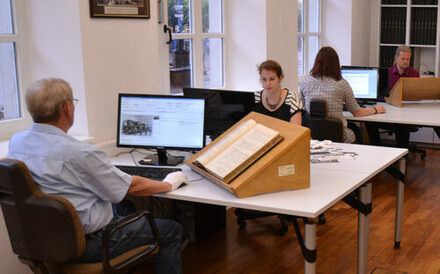 Das Bild zeigt Menschen, die im Lesesaal vor Recherche-PCs und historischen Dokumenten sitzen.